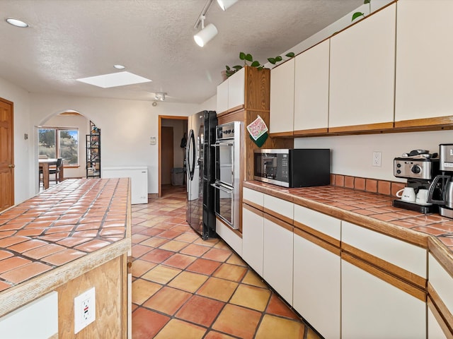 kitchen featuring stainless steel microwave, tile countertops, freestanding refrigerator, arched walkways, and white cabinetry