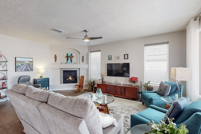 living room featuring visible vents, a ceiling fan, a textured ceiling, a glass covered fireplace, and dark wood finished floors