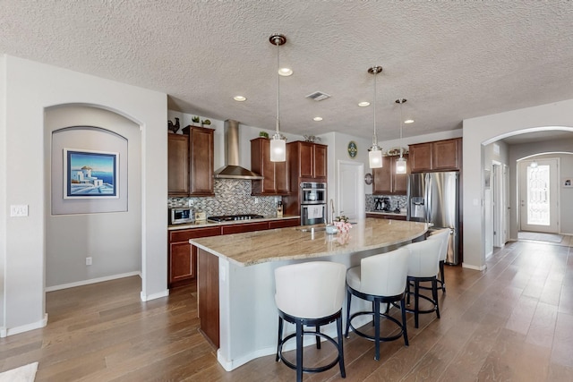 kitchen featuring a spacious island, backsplash, wall chimney range hood, arched walkways, and stainless steel appliances