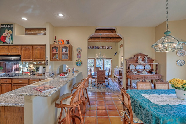 kitchen featuring range hood, arched walkways, brown cabinetry, a notable chandelier, and a sink