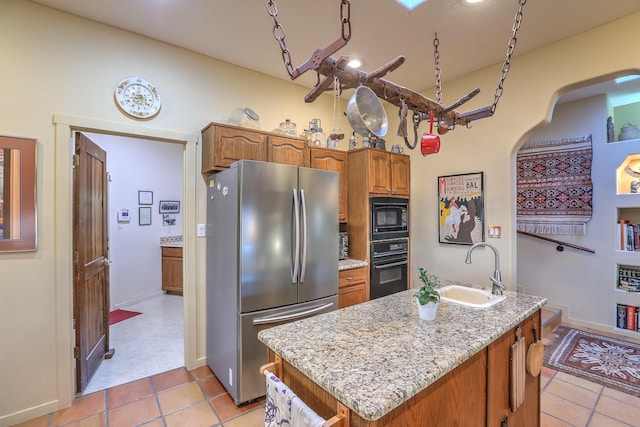 kitchen featuring a center island with sink, light stone counters, brown cabinets, black appliances, and a sink