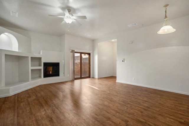 unfurnished living room featuring visible vents, built in shelves, wood finished floors, a glass covered fireplace, and a ceiling fan