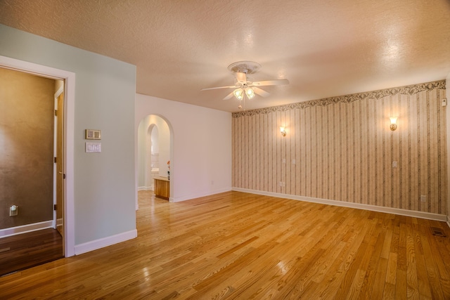 unfurnished room with a ceiling fan, baseboards, arched walkways, light wood-style floors, and a textured ceiling