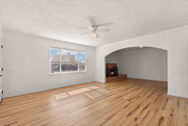 unfurnished living room with wood finished floors, a ceiling fan, arched walkways, a warm lit fireplace, and a textured ceiling