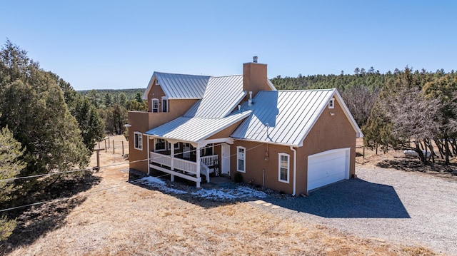 back of house featuring gravel driveway, a standing seam roof, a chimney, a garage, and metal roof