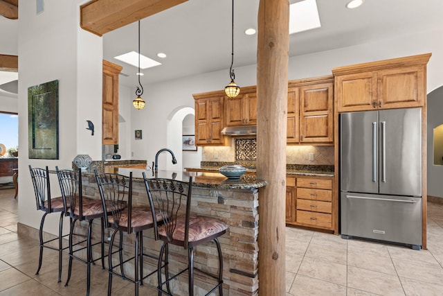 kitchen featuring a kitchen breakfast bar, dark stone countertops, high end fridge, light tile patterned flooring, and under cabinet range hood