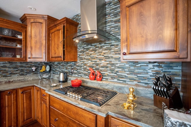 kitchen featuring backsplash, glass insert cabinets, wall chimney range hood, light stone counters, and stainless steel gas stovetop