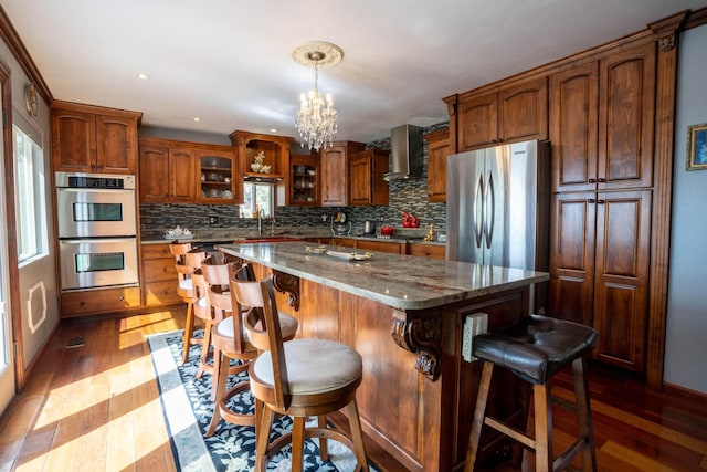 kitchen featuring decorative backsplash, glass insert cabinets, light wood-style floors, appliances with stainless steel finishes, and wall chimney range hood