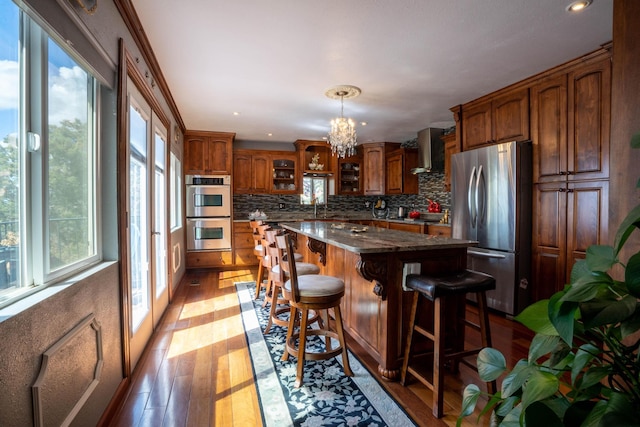 kitchen with light wood finished floors, a kitchen island, wall chimney range hood, a notable chandelier, and stainless steel appliances