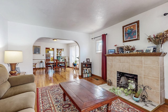 living area featuring arched walkways, light wood-style flooring, a tiled fireplace, and baseboards