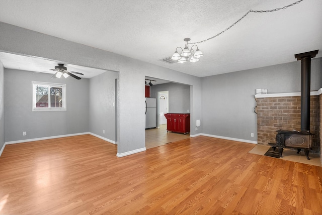 unfurnished living room with baseboards, light wood-style flooring, a wood stove, a textured ceiling, and ceiling fan with notable chandelier