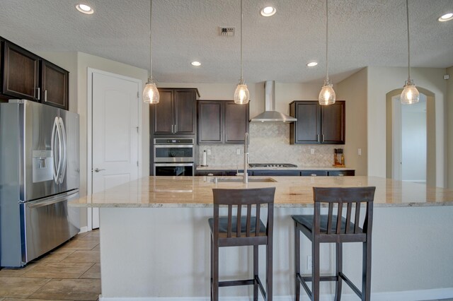 12136 Bear Valley Ln NW, Albuquerque, NM 87120 - kitchen with appliances with stainless steel finishes, wall chimney range hood, dark brown cabinetry, tasteful backsplash, and a textured ceiling