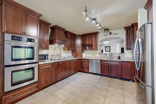 kitchen featuring decorative backsplash, a textured ceiling, appliances with stainless steel finishes, a sink, and premium range hood