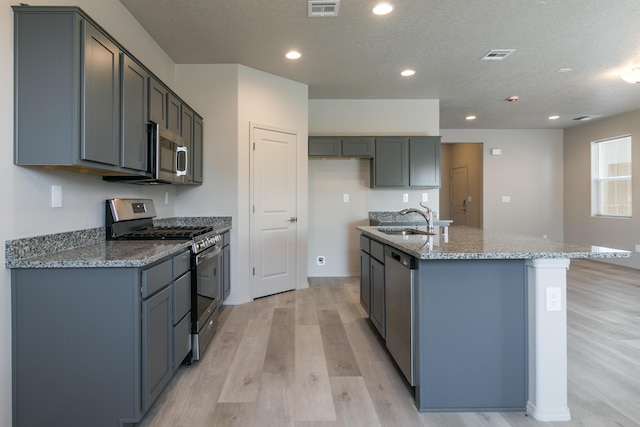 9600 Sage Ranch Ct SW, Albuquerque, NM 87121 - kitchen featuring appliances with stainless steel finishes, light wood finished floors, stone counters, a kitchen island with sink, and a textured ceiling