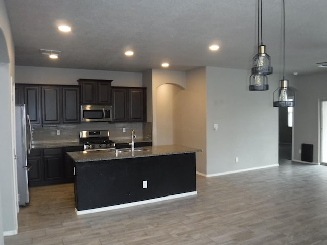 kitchen featuring an island with sink, stainless steel appliances, a sink, arched walkways, and tasteful backsplash