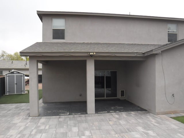 rear view of property featuring stucco siding, a patio area, a shingled roof, and a storage shed