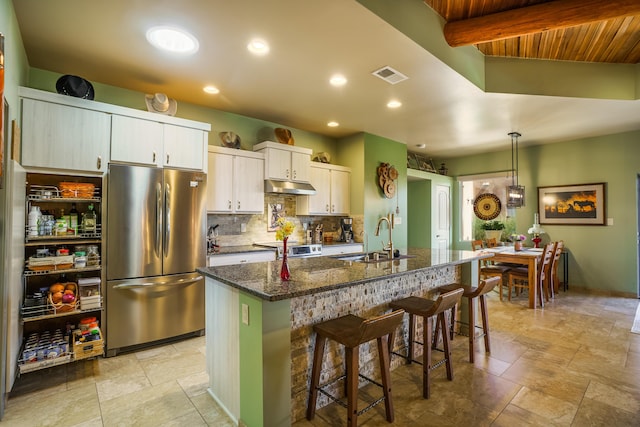kitchen with freestanding refrigerator, visible vents, under cabinet range hood, a sink, and a center island with sink