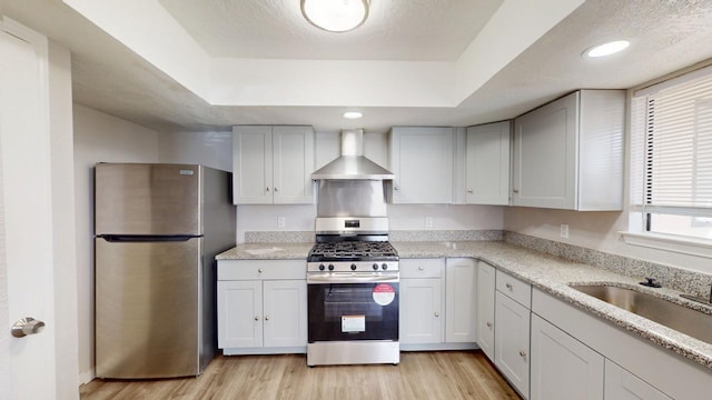 kitchen with a tray ceiling, light wood-type flooring, wall chimney range hood, and stainless steel appliances