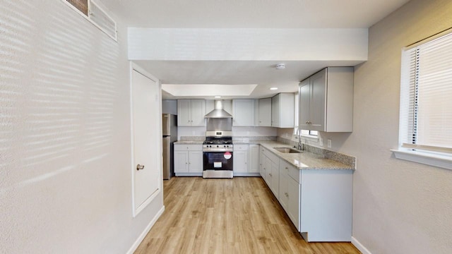 kitchen with light wood finished floors, light stone counters, stainless steel appliances, a sink, and wall chimney exhaust hood