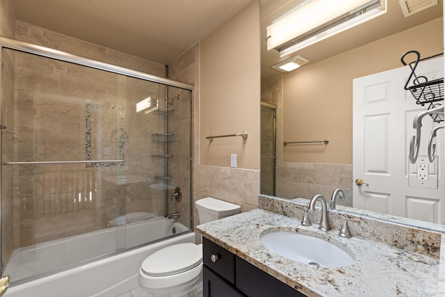 bathroom with tile walls, toilet, combined bath / shower with glass door, vanity, and wainscoting