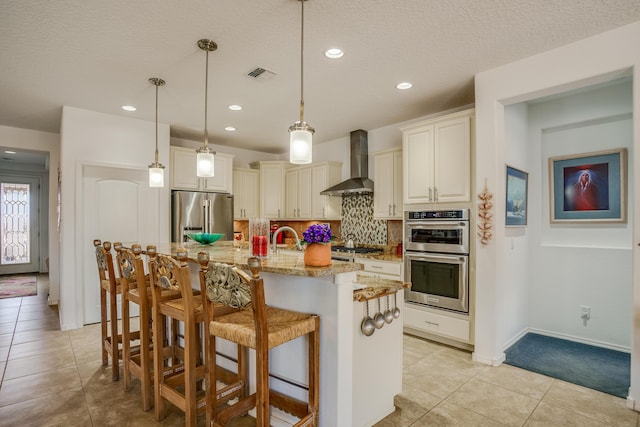 kitchen with tasteful backsplash, wall chimney range hood, light stone countertops, visible vents, and appliances with stainless steel finishes