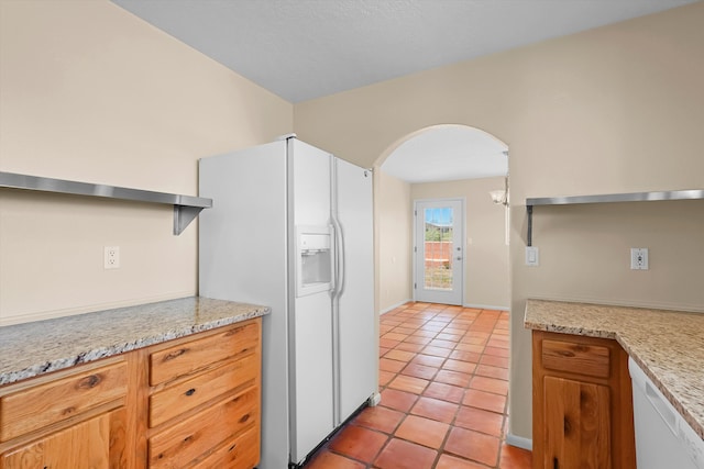 kitchen featuring brown cabinetry, arched walkways, light tile patterned floors, baseboards, and white appliances