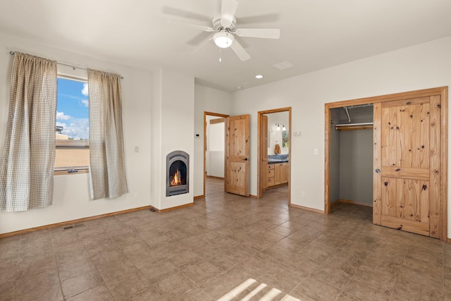 unfurnished bedroom featuring baseboards, a warm lit fireplace, a closet, ceiling fan, and ensuite bath