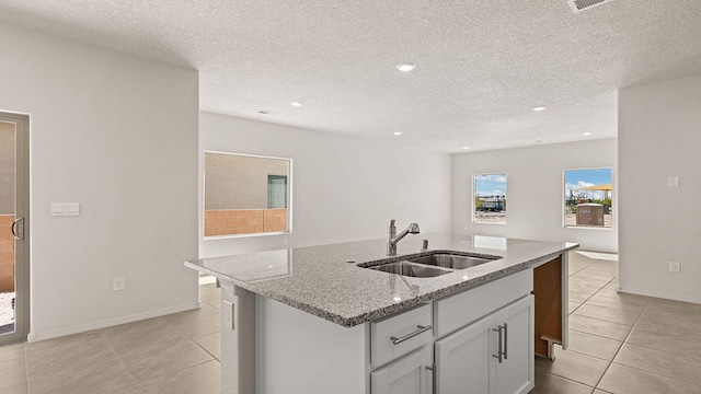 kitchen with light stone countertops, a sink, a textured ceiling, light tile patterned floors, and a center island with sink