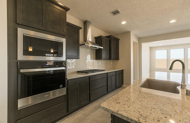 kitchen with a sink, light stone counters, visible vents, wall chimney exhaust hood, and appliances with stainless steel finishes