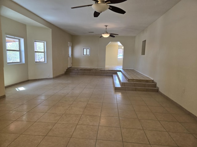 empty room with arched walkways, a ceiling fan, light tile patterned flooring, and baseboards