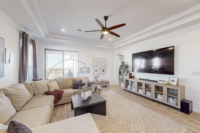 living room with ceiling fan, arched walkways, light wood finished floors, a raised ceiling, and visible vents