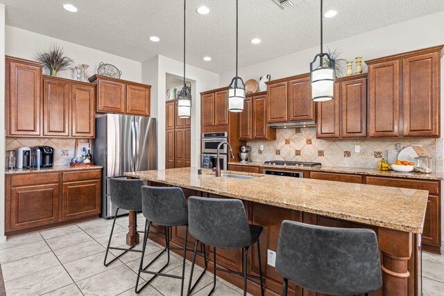 4024 Colina Roja Ln NE, Rio Rancho, NM 87124 - kitchen featuring stainless steel appliances, brown cabinetry, an island with sink, a textured ceiling, and backsplash