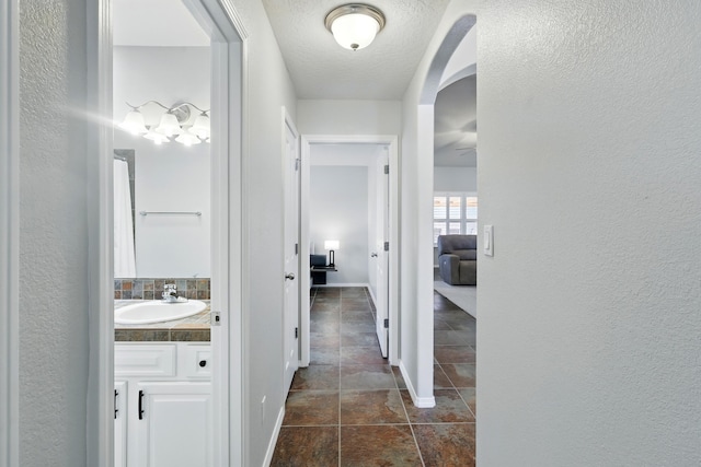 hallway with a sink, a textured wall, a textured ceiling, baseboards, and arched walkways