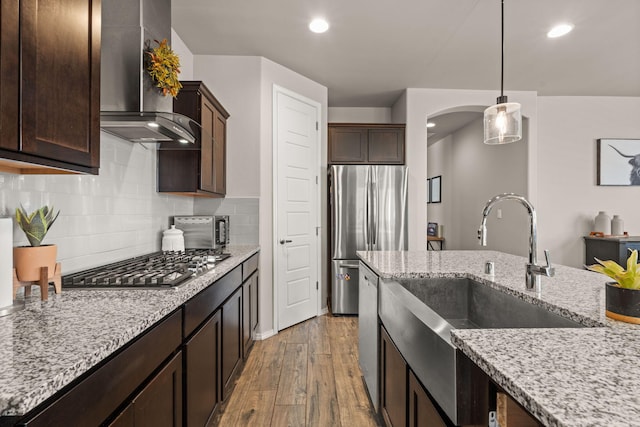 kitchen with a sink, stainless steel appliances, wall chimney range hood, arched walkways, and dark brown cabinets