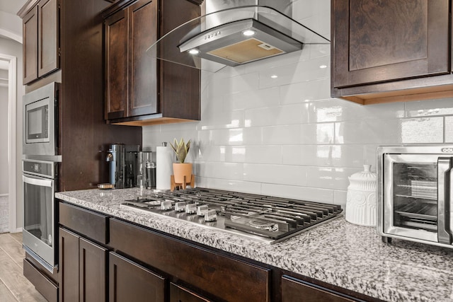 kitchen featuring wall chimney range hood, light stone counters, appliances with stainless steel finishes, dark brown cabinets, and backsplash