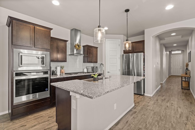 kitchen with a sink, appliances with stainless steel finishes, dark brown cabinets, light wood finished floors, and wall chimney exhaust hood