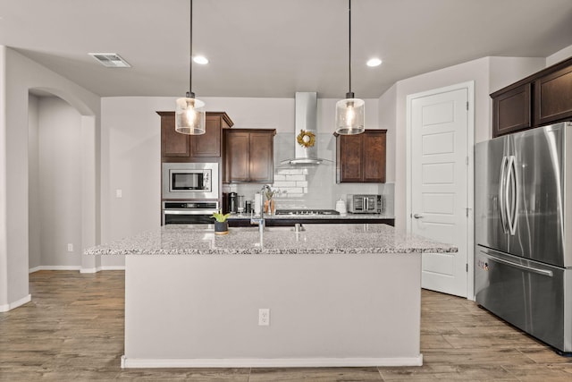 kitchen featuring light stone counters, wall chimney range hood, tasteful backsplash, appliances with stainless steel finishes, and dark brown cabinets