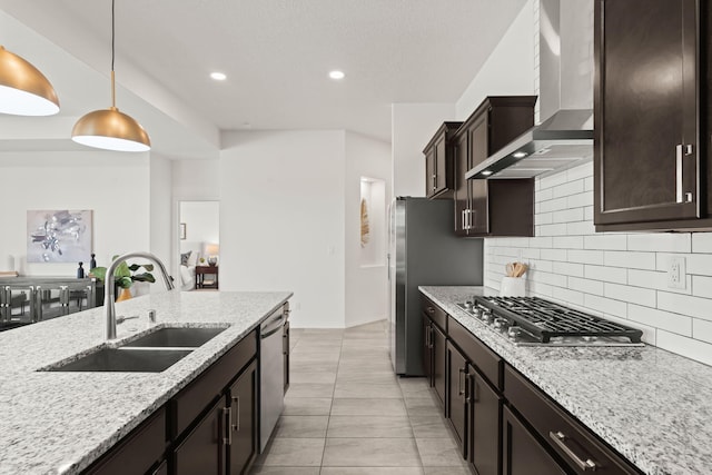 kitchen featuring wall chimney range hood, light stone counters, a sink, backsplash, and stainless steel appliances
