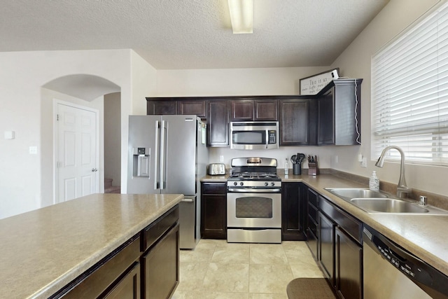 kitchen featuring arched walkways, stainless steel appliances, dark brown cabinets, a sink, and a textured ceiling