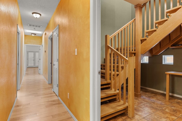corridor with visible vents, light wood-type flooring, baseboards, a textured wall, and stairs
