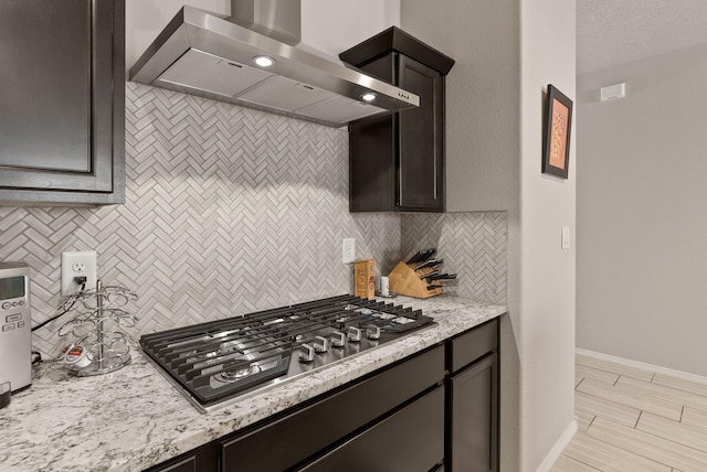 kitchen with wall chimney range hood, light stone countertops, stainless steel gas cooktop, baseboards, and backsplash