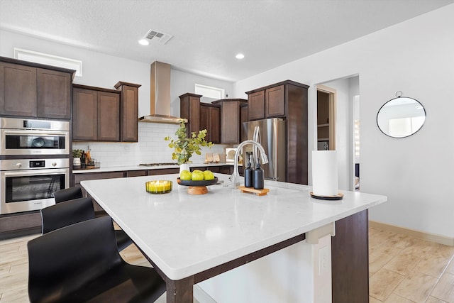 kitchen with dark brown cabinetry, visible vents, appliances with stainless steel finishes, wall chimney exhaust hood, and an island with sink