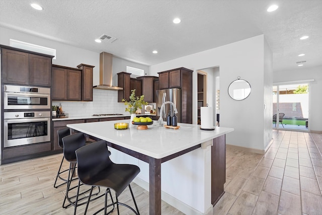 kitchen featuring wall chimney range hood, appliances with stainless steel finishes, a textured ceiling, visible vents, and a center island with sink