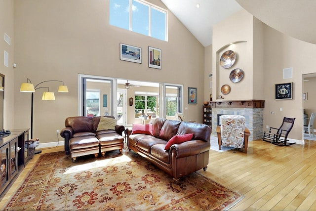 living area with high vaulted ceiling, light wood finished floors, baseboards, a stone fireplace, and a ceiling fan