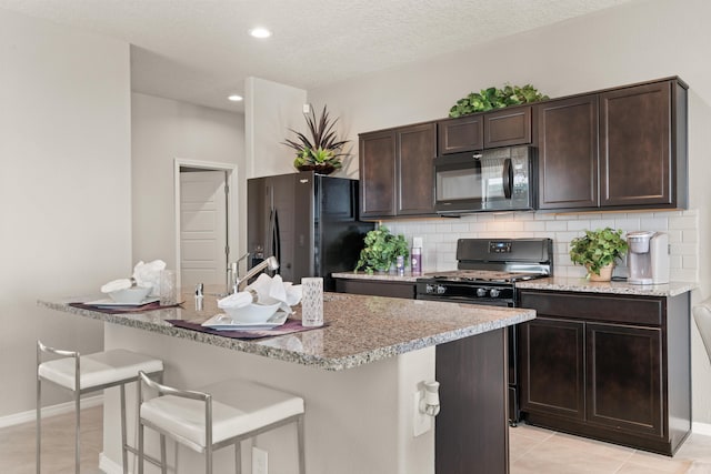 kitchen featuring black appliances, light tile patterned floors, backsplash, dark brown cabinets, and an island with sink