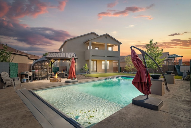 pool at dusk with an outdoor fire pit, a patio, fence, and a gazebo