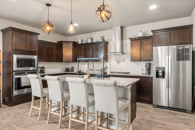 kitchen with appliances with stainless steel finishes, wall chimney exhaust hood, dark brown cabinetry, light stone countertops, and backsplash