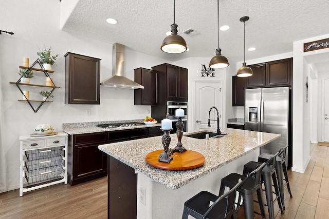 11805 Badger Mountain Trl NW, Albuquerque, NM 87120 - kitchen with wall chimney range hood, stainless steel appliances, light wood finished floors, dark brown cabinets, and a textured ceiling