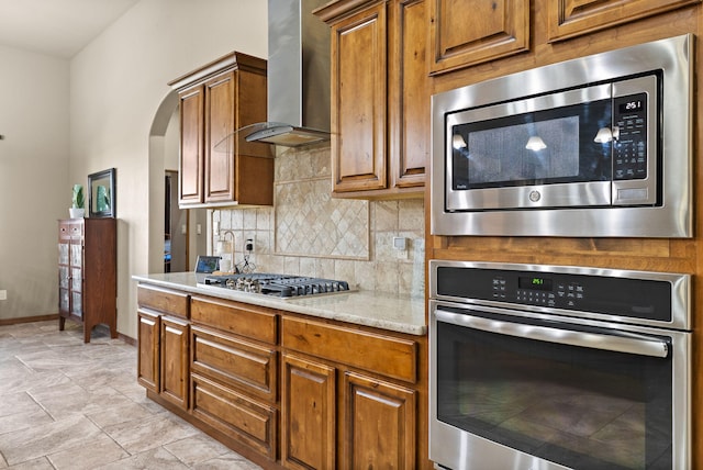 9844 Cameron St NW, Albuquerque, NM 87114 - kitchen with stainless steel appliances, wall chimney exhaust hood, decorative backsplash, arched walkways, and brown cabinetry