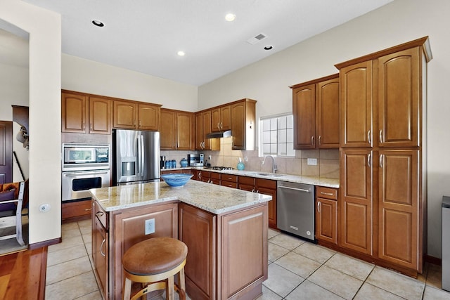 8712 Sandwater Rd NW, Albuquerque, NM 87120 - kitchen featuring appliances with stainless steel finishes, light stone countertops, a center island, backsplash, and brown cabinetry